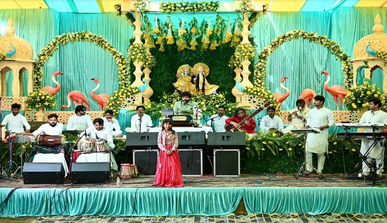 RV Band performing a seated sankirtan on a garden-themed stage