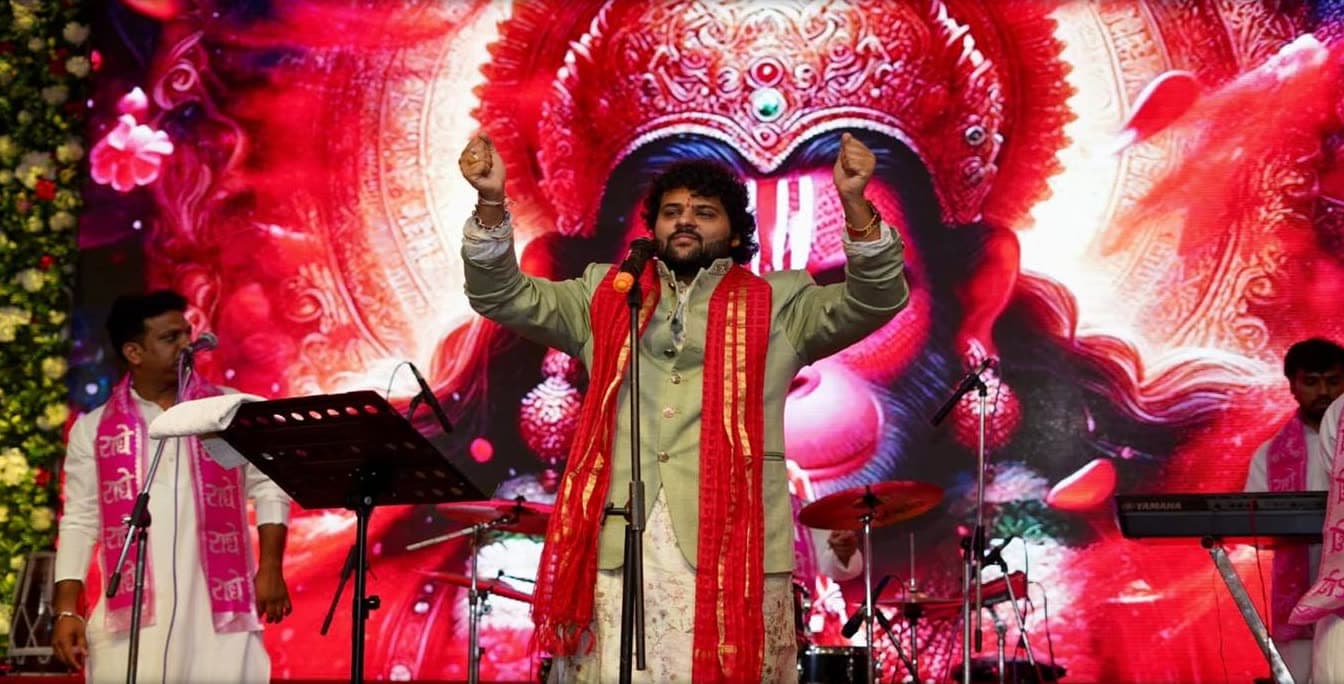 Pt. Govinda Sharma singing with both arms raised in front of a large Hanuman ji backdrop